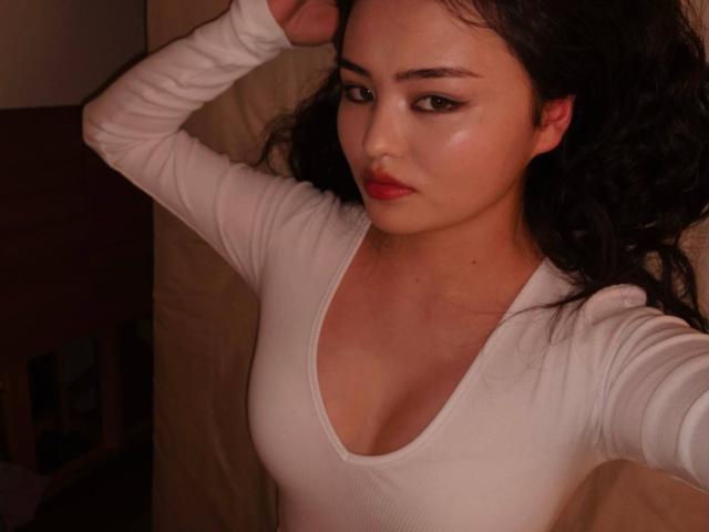Sovelle has curly black hair, red lipstick, and a white, long-sleeve, V-neck top showing her small, firm tits. She's looking at the camera with a serious expression.