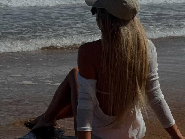 MissiSofiya with long blonde hair, white off-shoulder top, and cap, sitting on sandy beach, facing ocean, legs bent, ass slightly visible, wearing sandals, back to camera.