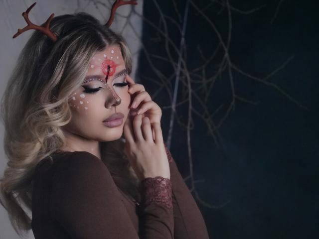FollowLove with blonde hair, deer antlers, and white facial paint dots, wearing a brown top, hands near face, looking down, soft light, dark background, wavy hair.