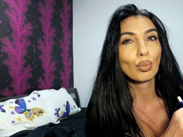 BrunetteBabe69 with long black hair, brown eyes, and pouty lips, holding a makeup brush near her face, wearing a white floral pillow in the background.
