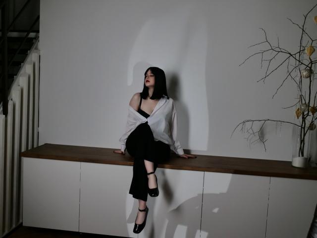 HoneyGlow with black hair, red lipstick, white off-shoulder shirt, black pants, and black heels, sitting on a wooden bench, legs crossed, shadow on white wall.