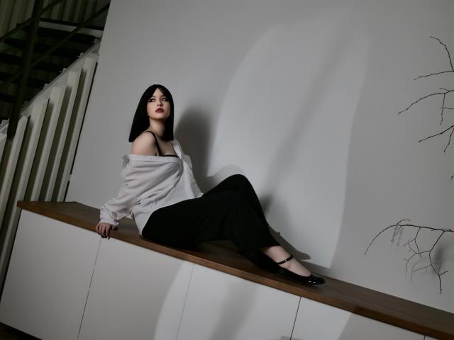 HoneyGlow with black hair, off-shoulder white shirt, black pants, and black flats, sitting on a wooden shelf, exposing her tits, with a shadow on a white wall.