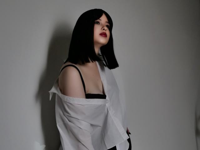 HoneyGlow with black bob, red lipstick, off-shoulder white shirt, black bra straps visible, looking up, shadow on gray wall, subtle smile, sexy, confident, soft lighting.