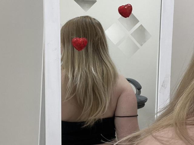 DiLaria's blote rug en lange blonde haar voor een spiegel, met rode hartstickers die haar borsten bedekken. Ze draagt een zwarte top zonder bandjes en een zwarse armband..
