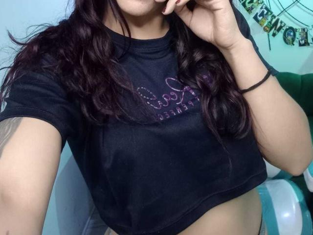 AriannaSeete with dark wavy hair, wearing a black crop top, showing her bare midriff, and touching her face, her hand has a black bracelet.