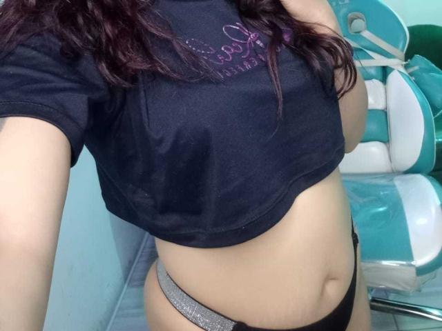 AriannaSeete's midriff with her black t-shirt lifted, showing her small tits and black thong with gray glitter. She has wavy dark hair, pale skin, and a slight belly.