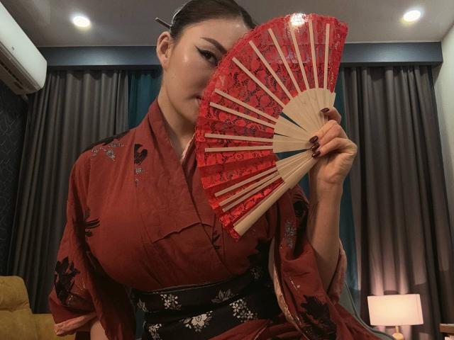 PlumHaze with dark hair in a red floral kimono, holding a red lace fan, showing off her tits, in a dimly lit room with gray curtains.
