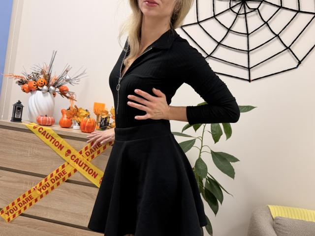 Rorria porte une robe noire à fermeture éclair avec une jupe évasée, une main sur la hanche, et se tient à côté d'une commode en bois décorée pour Halloween, avec du ruban adhésif jaune de signalisation..
