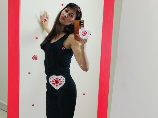 MiraMixi with long brown hair, black tank top, and black skirt, showing a heart with red roses on her butt in a mirror selfie, holding an orange phone.
