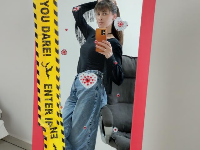 MiraMixi with tits covered in white heart-shaped stickers, wearing a black top, blue baggy jeans, black cardigan, taking a mirror selfie with a orange phone.