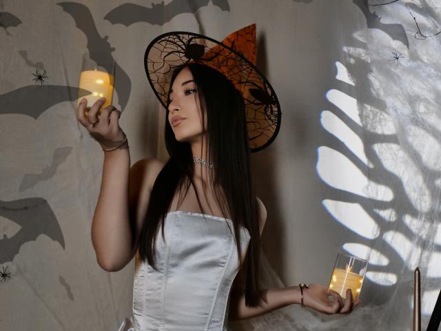 ElviraWesdety with long black hair, wearing a witch hat and white strapless corset, holding two candles, looking at the left candle, showing off her tits, in a spooky setting.