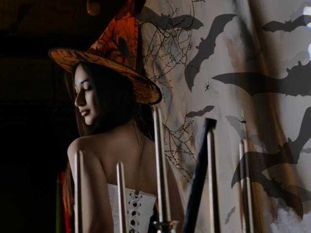 ElviraWesdety with dark hair, wearing a witch hat and strapless white top, showing her bare back and small ass, standing in a dimly lit room with candles.