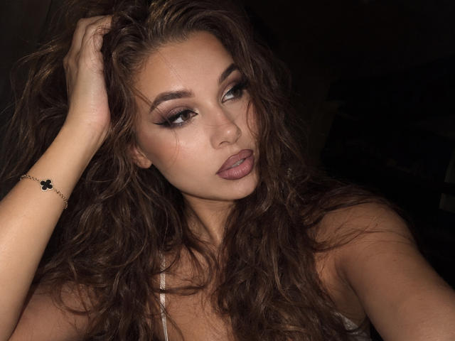 Niferttiti with wavy brown hair, pouty lips, and smoky eye makeup, touching her hair, wearing a thin bracelet, looking seductively at the camera. Dark background.