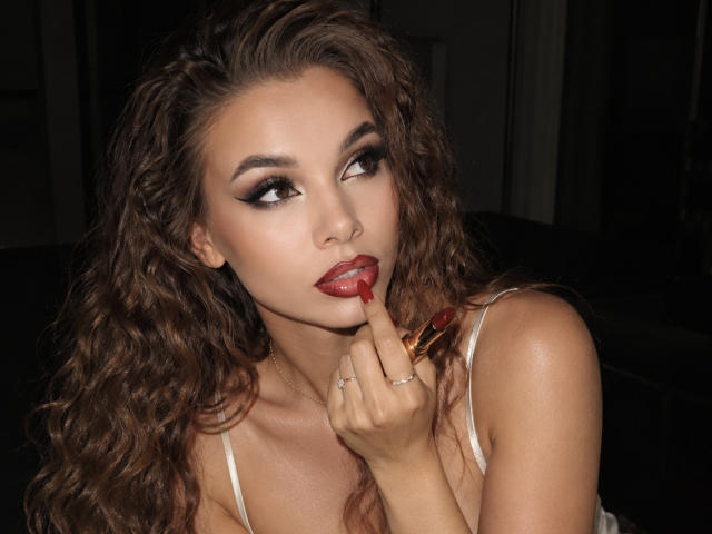 Niferttiti with curly brown hair, red lipstick, and smoky eye makeup, sucking on a red lipstick with finger, wearing a white spaghetti strap top, looking up seductively.