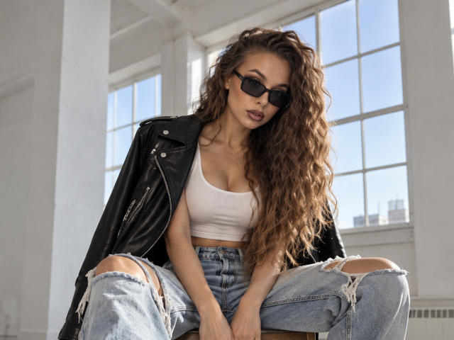 Niferttiti with long curly brown hair, wearing black sunglasses, white crop top, black leather jacket, and ripped jeans, sitting in bright room, showcasing her big boobs and denim ass.