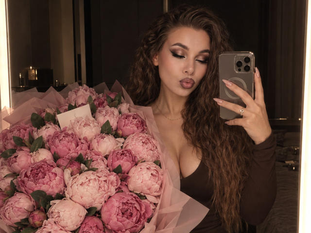 Niferttiti with long curly brown hair, brown top, big boobs, holding pink roses, tongue out, taking mirror selfie with iPhone, ring on finger, dark room, candles in background.