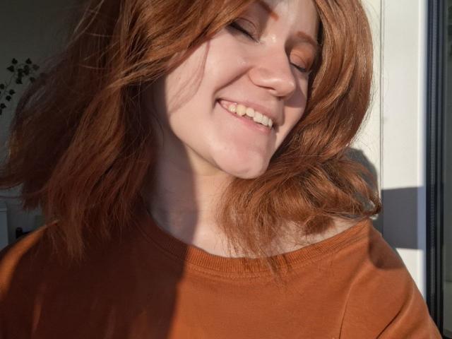 Charissa Lathem, con i capelli rossi, sorridente, con gli occhi chiusi, indossa una maglietta arancione, la luce del sole proietta ombre sul suo viso, capelli leggermente ondulati, trucco naturale, sorriso dolce, espressione rilassata..