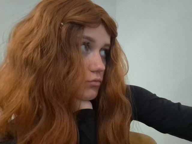 Charissa Lathem ha i capelli rossi e ondulati che le coprono il viso, indossa una maglia nera e ha un'espressione seria, appoggiando il mento sulla mano. I suoi occhi azzurri guardano di lato..
