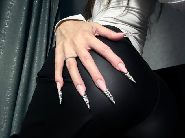 AngelesHazim's hand with long, sharp, black-and-white patterned nails rests on her black ass, wearing a white top, ring on finger, against a grey curtain.