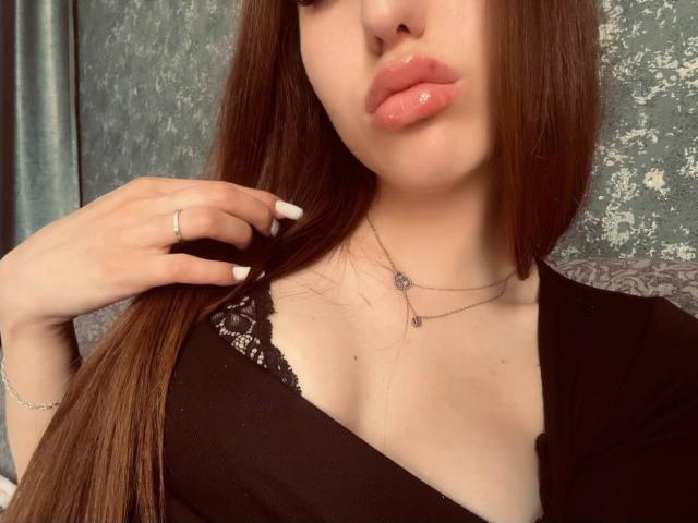 AngelesHazim with glossy pink lips, long brown hair, black lace top, holding hair with white nails, silver necklace, ring, and bracelet, showing her tits.