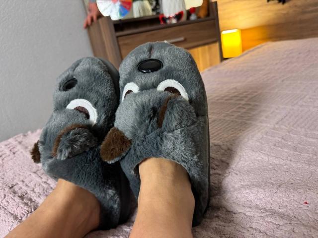 Niferttiti's legs in fluffy gray bear slippers with white eyes and brown noses, on a pink textured bed. Slippers cover their feet, showing only their lower legs.
