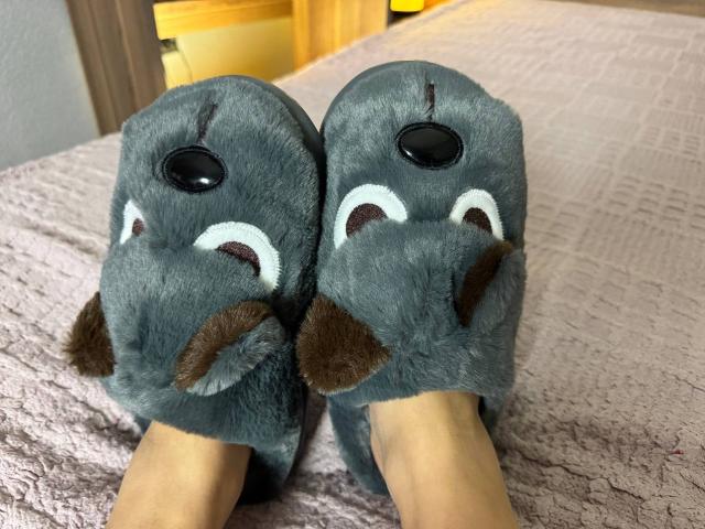 Niferttiti's feet are in fuzzy, gray, plush slippers with black noses and white, brown, and dark brown eyes, lying on a pink textured bedspread.