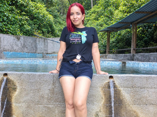 KateMonroe, avec des cheveux roux, un t-shirt noir et un short en jean, appuyée contre une fontaine en béton, de l'eau qui goutte, un tatouage sur le bras droit, souriante, cheveux courts, tenue décontractée, environnement extérieur..