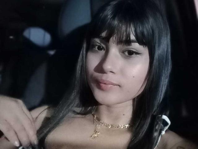 AshlyLee has dark straight hair, bangs, and a gold necklace with a small pendant. She's smiling slightly, showing subtle makeup and a small mole on her right cheek.