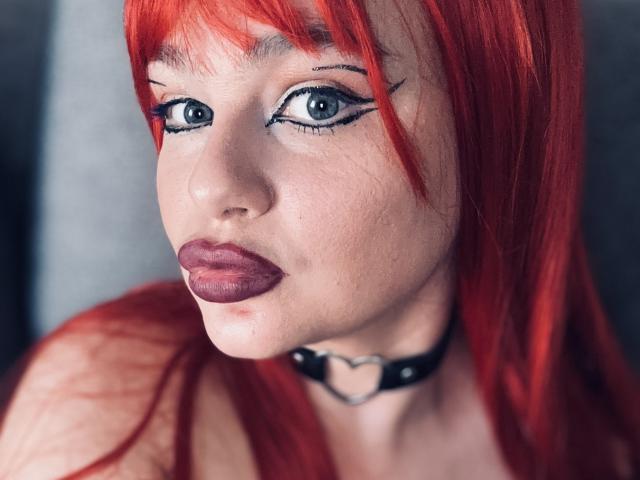 LunaOreilly has red hair, blue eyes with black eyeliner, purple lipstick, a black choker with a heart, and pale skin. She's looking directly at the camera with a pouty face.