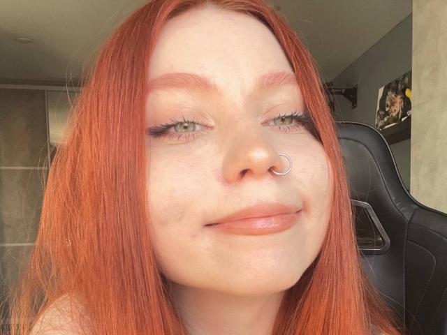 LilliamChevalier with red hair, nose ring, and slight smile, has light freckles and wears subtle makeup with long eyelashes, looking directly at the camera.