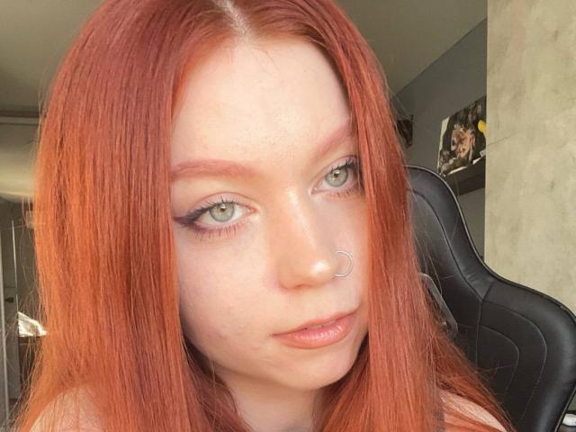 LilliamChevalier has red hair, green eyes, and a nose ring. Her makeup includes black eyeliner and pinkish lipstick. She's sitting in a black chair, looking at the camera.