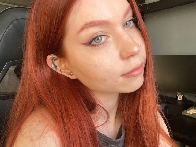 LilliamChevalier has red hair, green eyes, and a nose ring. She's wearing a gray top, with subtle makeup and a slight smile, looking directly at the camera.