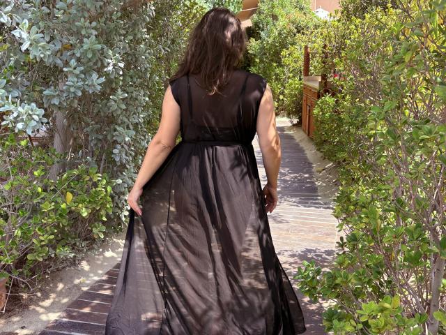 BadAngel with long brown hair, black sheer dress, and visible ass, walking on a wooden path, surrounded by green bushes. Her dress flutters slightly with her movement.