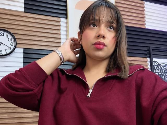 ZaraBlaze with brown hair, red zip-up hoodie, pink lipstick, and silver bracelet, hand in hair, looking at camera. Her ass is not visible. Clock on wooden wall.
