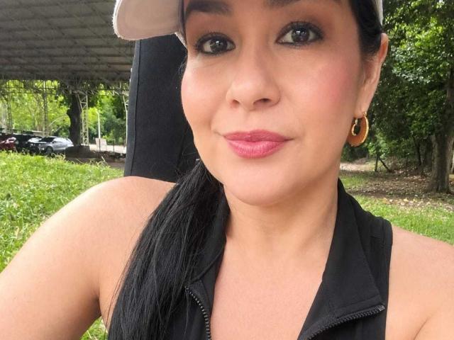 TamaraFoxy, con i capelli neri raccolti in una coda di cavallo, indossa una canotta nera, orecchini a cerchio dorati, rossetto rosa e un berretto beige, sorridendo alla fotocamera..