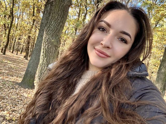 AquaLady with wavy brown hair, full lips, and dark eyebrows, wearing a black puffer jacket, smiling softly, standing in a forest with sunlit leaves.