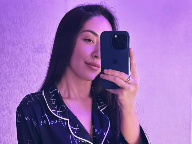 AquaLady with long dark hair, wearing a black silk robe with white patterns, holding a blue iPhone, taking a selfie with purple lighting, smiling softly.