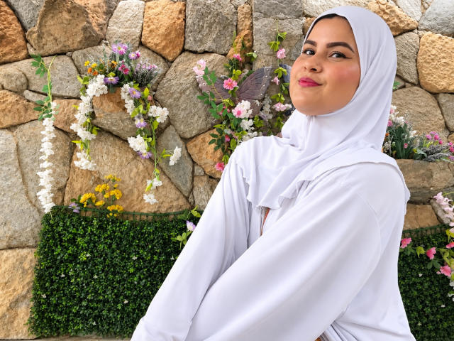 AishaAmin with a white hijab and long-sleeve white dress, smiling at the camera, showing off her ass, with pink and white flowers in the background.