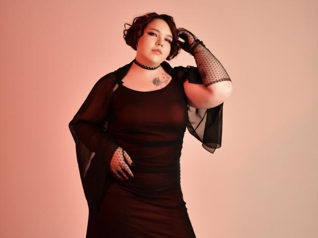 IvoryeFlame in a sheer black dress, polka dot gloves, choker, and short curly brown hair, showing tattoos, hand on hip, looking confident with a sexy, full-ass figure.