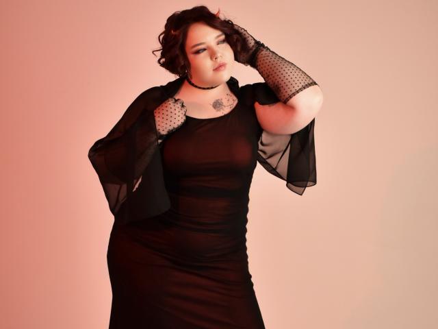 IvoryeFlame, chubby with big tits, wearing a sheer black dress, black polka dot gloves, and choker, standing with hands on shoulders, looking down, against pink background.