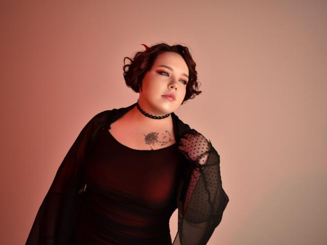 IvoryeFlame with curly brown hair, wearing a black sheer dress, polka dot sleeves, choker, and tattoo on chest, has a sultry expression, and a slight downward gaze.