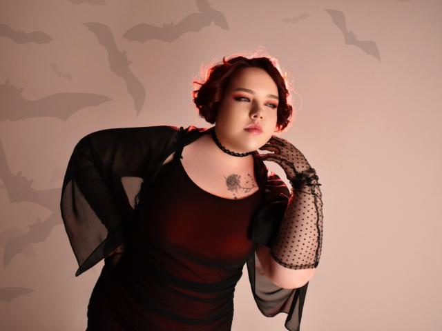 IvoryeFlame with red curly hair, black sheer dress, polka dot gloves, choker, and tattoo, standing confidently, hand on cheek, looking sexy, bats shadow in background.
