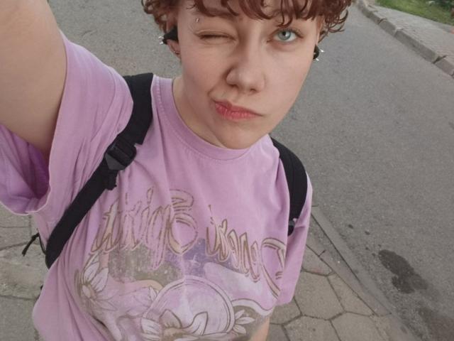 PhilomenaDeni with short curly brown hair, winking, wearing a purple "Dreaming" t-shirt, black backpack, and red lipstick, taking a selfie outdoors on a paved street.