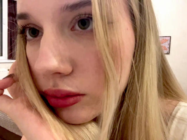 MayeAlberry tiene la piel clara, cabello rubio, ojos verdes y lápiz labial rojo. Su mano está cerca de su rostro, con una expresión ligeramente fruncida. Lleva una blusa color beige..