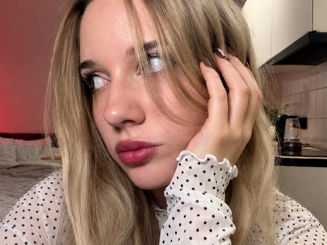 Het gezicht van MayeAlberry, met blond haar, roze lippen en een stippenblouse, kijkt opzij, met een hand op haar wang, glanzende nagels, in een keuken met rood licht..