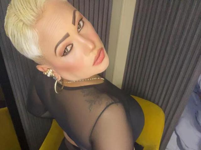 Gianella Cox, avec ses cheveux blonds courts, regardant par-dessus son épaule, portant un haut noir transparent, des boucles d'oreilles cerceaux et du rouge à lèvres rose, montrant ses tatouages et une chaise jaune dans la photo..
