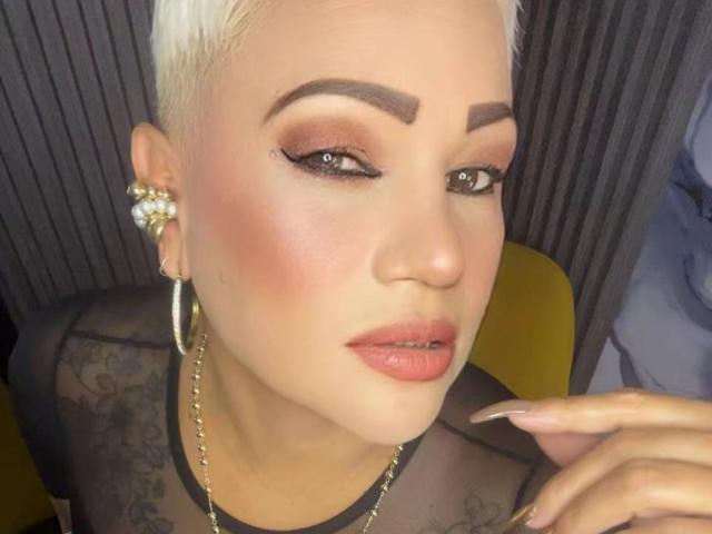 Gianella Cox a les cheveux courts, d'un blond platine, des sourcils épais et du rouge à lèvres rose. Elle porte de grandes boucles d'oreilles et un haut noir transparent, avec un collier en chaîne dorée..