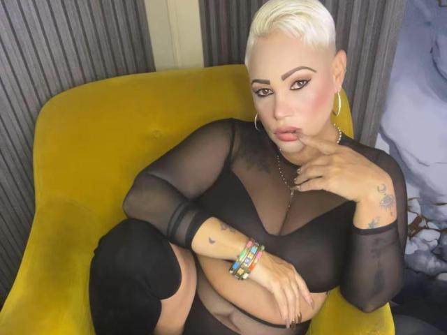Gianella Cox, avec ses cheveux blonds courts, ses boucles d'oreilles, son haut noir transparent, ses bas noirs montants jusqu'à la cuisse et ses bracelets colorés, faisant une moue avec la main près de la bouche, assise sur une chaise jaune..