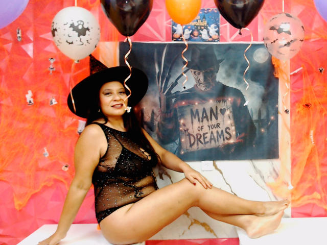 MiaCallie in a black witch hat, black see-through lingerie with sparkles, sitting with legs crossed, smiling, surrounded by balloons, and a "Man of Your Dreams" poster.