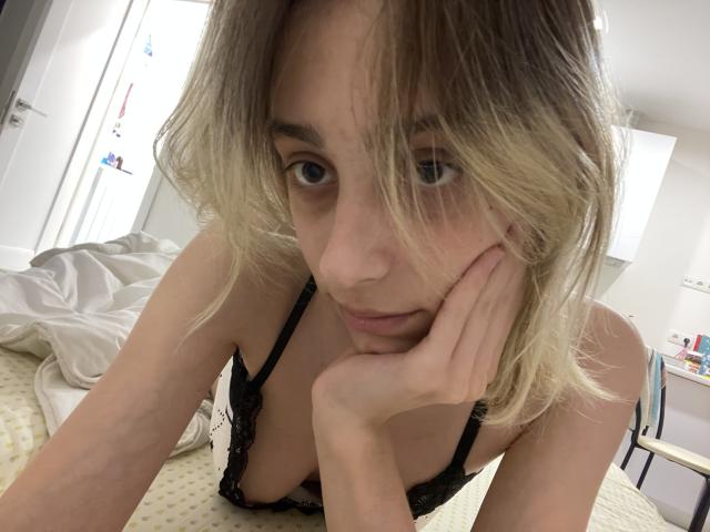 MilaGlow with blonde, wavy hair, looking up, has her hand on her cheek, wearing black lace lingerie, exposing her small tits, in a bedroom with a white bed.